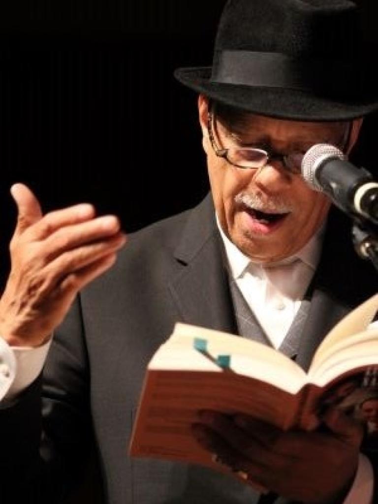 Dr. Haki R. Madhubuti reads from a book into a microphone, wearing a black suit and a black hat.