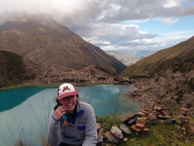 Photo of Linsay Carrera wearing a U of MN cap sitting by Lake Humantay in Southern Peru in spring 2019.