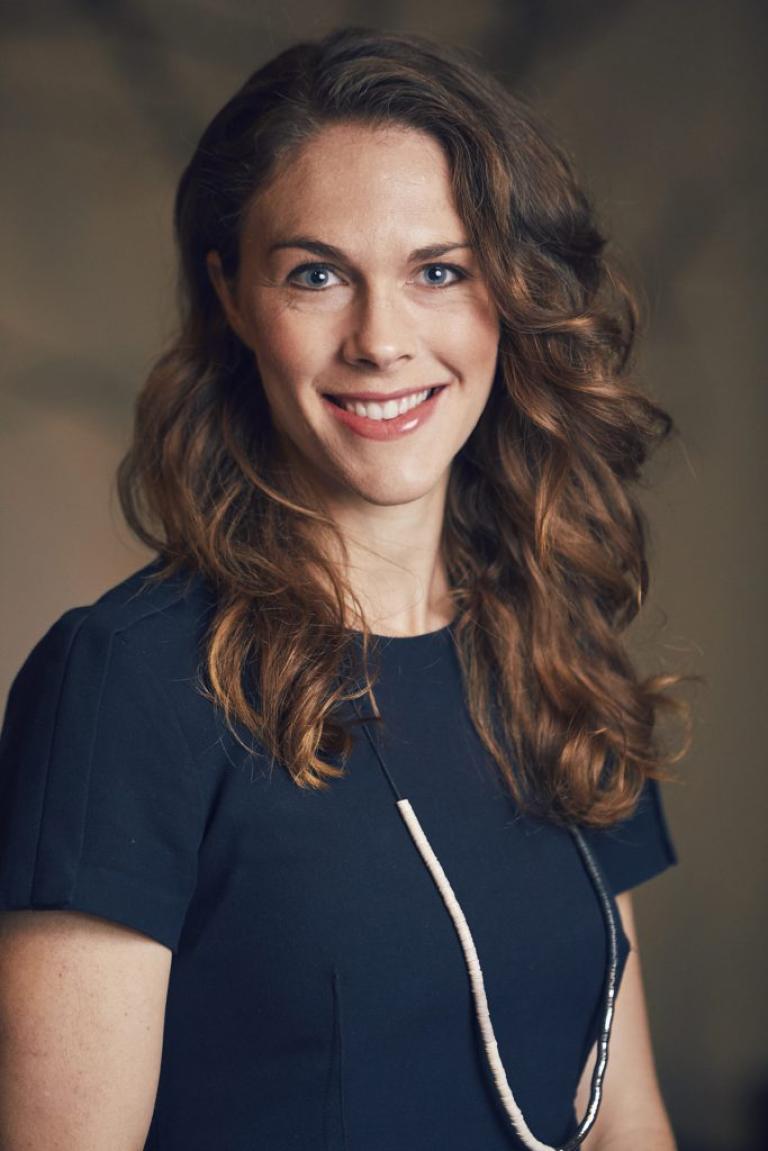 A headshot of Dr. Marti DeLiema. She wears a short-sleeved navy top, and her wavy hair down. She is smiling.