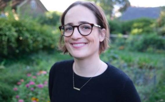 SSW PhD Student Bonnie Horgos smiles in front of a yard filled with flowers. She is wearing glasses, a dark shirt and a small necklace.