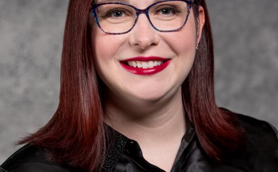 Head shot of Stephanie Hanson, PhD student. Stephanie has dark red hair, pale skin, black glasses and is wearing a shiny black shirt.