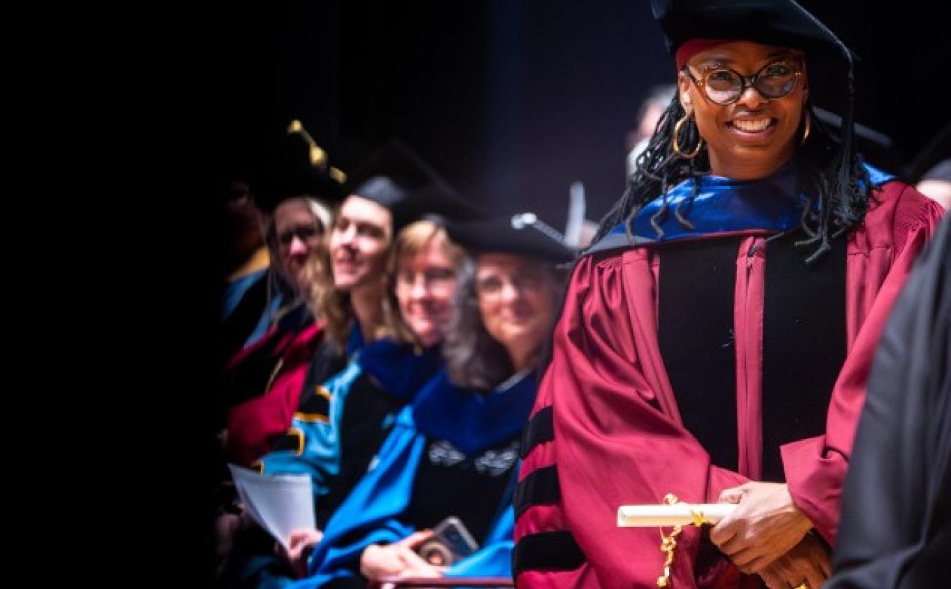 SSW faculty onstage at the 2024 School of Social Work graduation ceremony await graduates onstage, at the right of the image, standing in red ceremonial robes is Joan Blakey, PhD, director of the SSW.