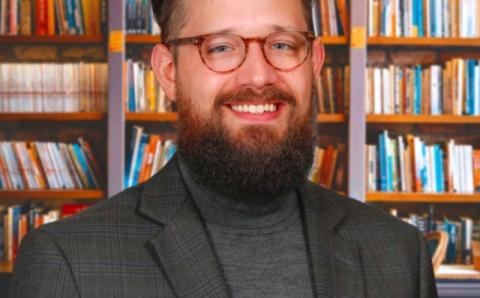 PhD candidate Jacob Otis smiles in a professional portrait. He has a beard and wears glasses, and stands in front of a full bookcase.