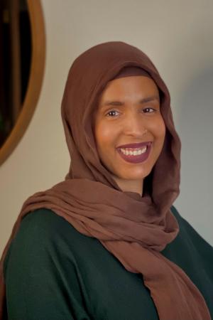 Johara Suileman, PhD candidate, wears a brown hijab and a green dress and smiles in this professional portrait.