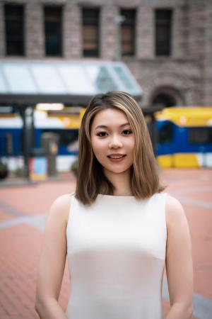 Louise (Siyu) Gao, PhD candidate, wears a white sleeveless dress and smiles in this professional portrait taken outdoors at the Saint Paul Farmer's Market with the light rail in the background.