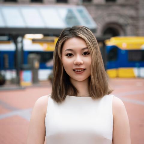 Louise (Siyu) Gao, PhD candidate, wears a white sleeveless dress and smiles in this professional portrait taken outdoors at the Saint Paul Farmer's Market with the light rail in the background.
